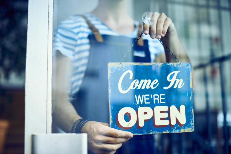 작업복을 입은 인물이 상점 창문에 "Come in, we're open"이라고 적힌 파란색 표지판을 들고 있습니다